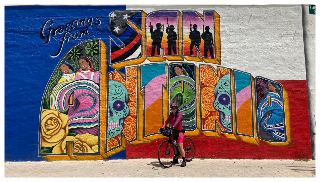 Ana Guajardo posing with her bike in front of a colorful mural that reads "San Antonio"