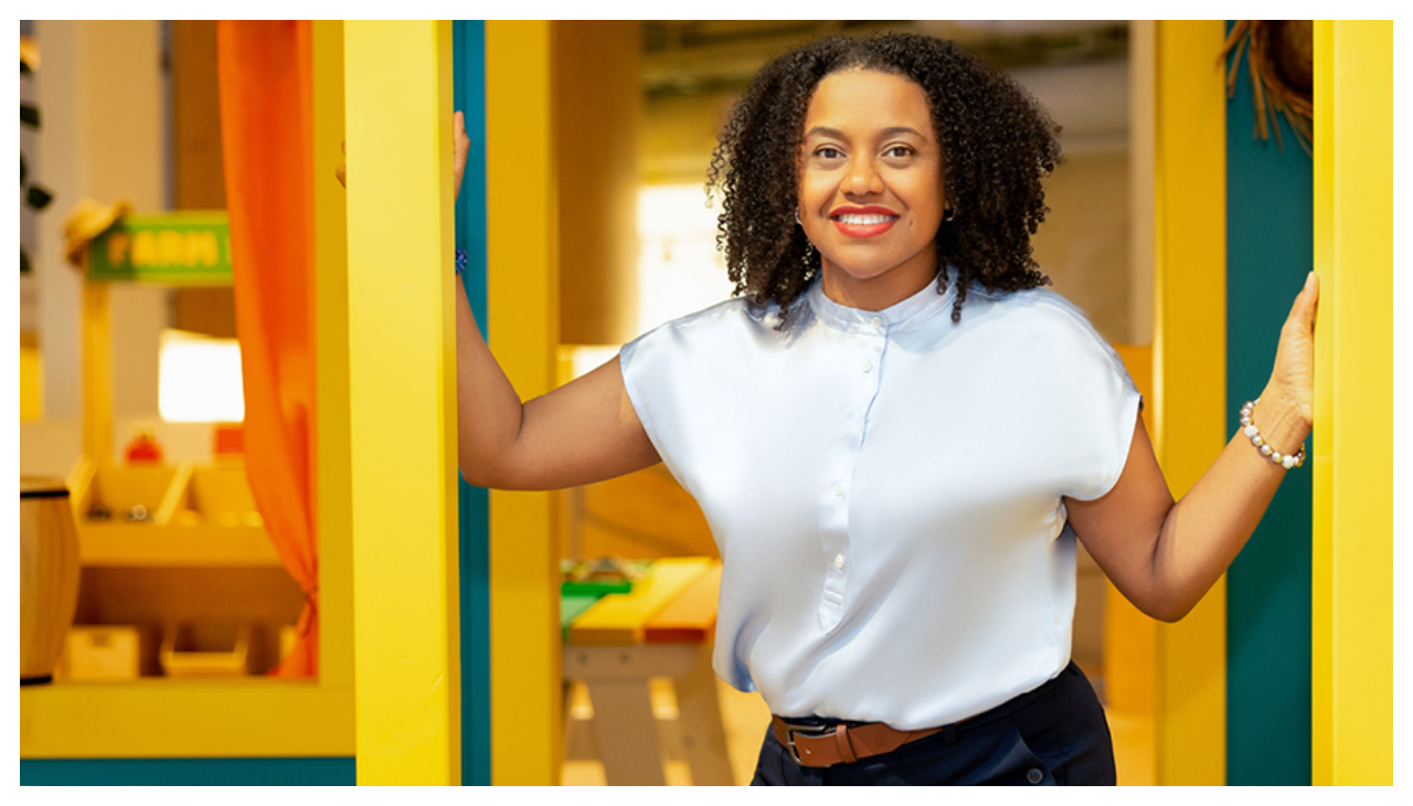 Denise Rosario Adusei, an Afro-Latina woman. She is wearing a light blue button up shirt and is standing in the doorway of a colorful room decorated for children. Her hands rest on each side of a doorway, and she is smiling towards the viewer.