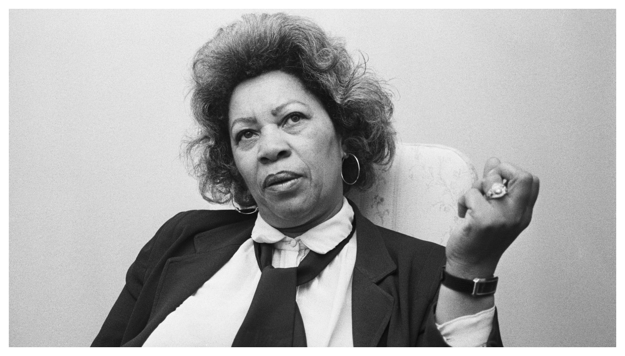 A black and white photo of Toni Morrison from the chest up. She is sitting down with one hand raised beside her, with a contemplative look on her face.