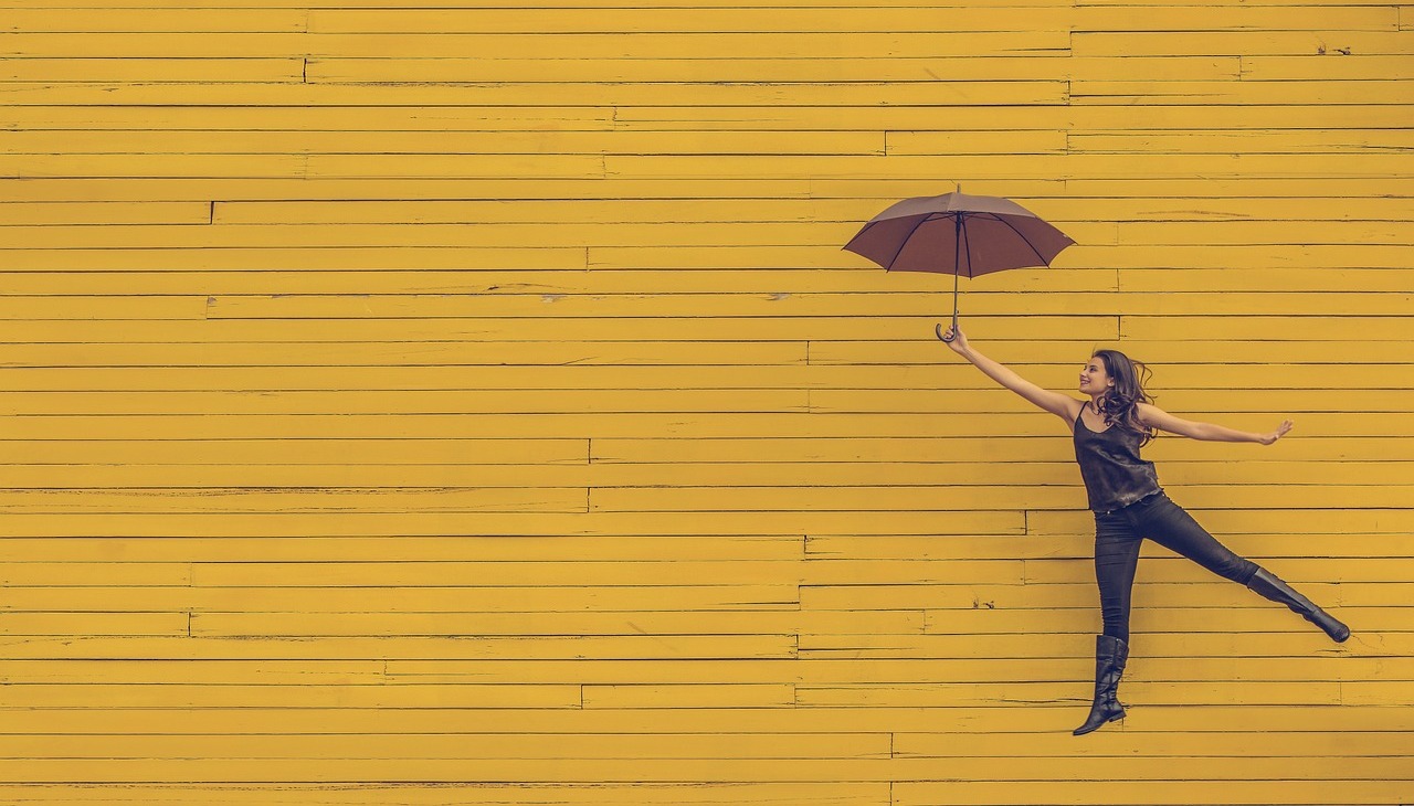 Woman jumps holding an umbrella next to a big yellow wall.
