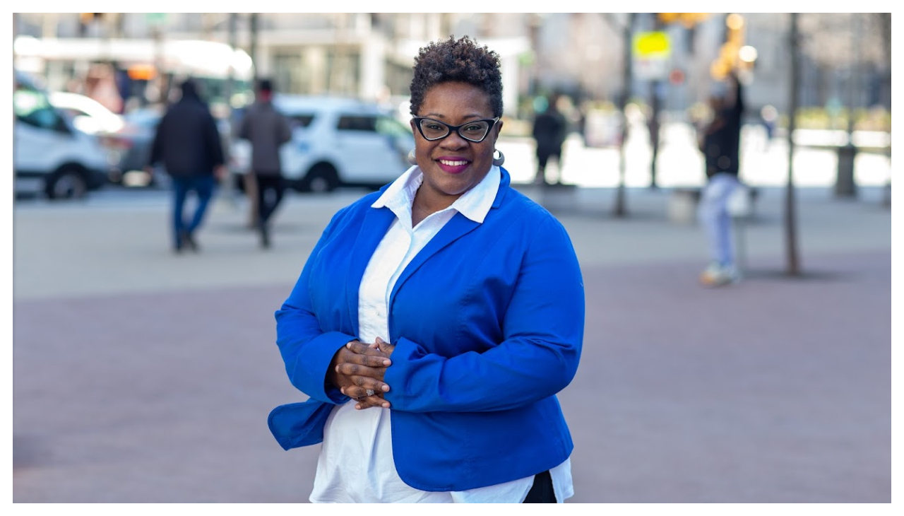 Reverend Naomi Washington-Leapheart standing in a public square. She is a Black woman wearing a blue coat with a pale blue button up shirt underneath it. She has short brown heair and is facing the viewer with her hands folded over her stomach. She is smiling.