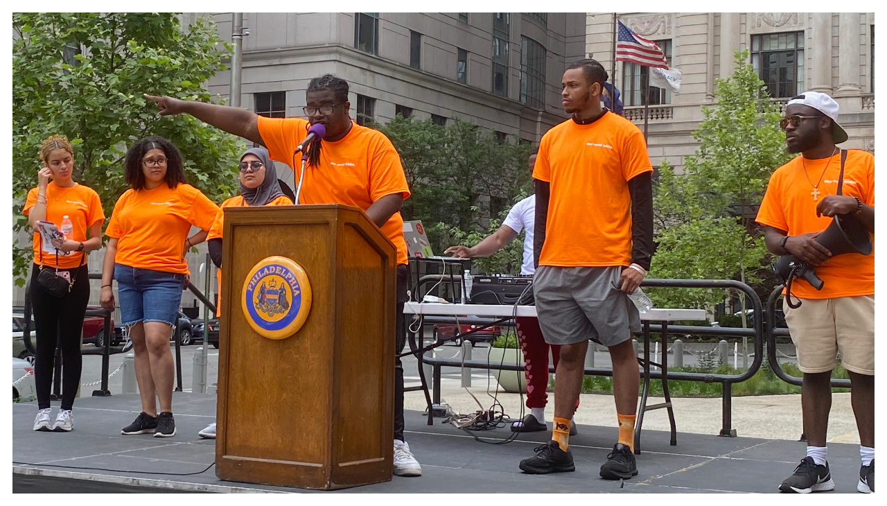 Several members of the Youth Unity Walk on a stage, listening to Jeron Williams II speak at a podium beside City Hall in Philadelphia.