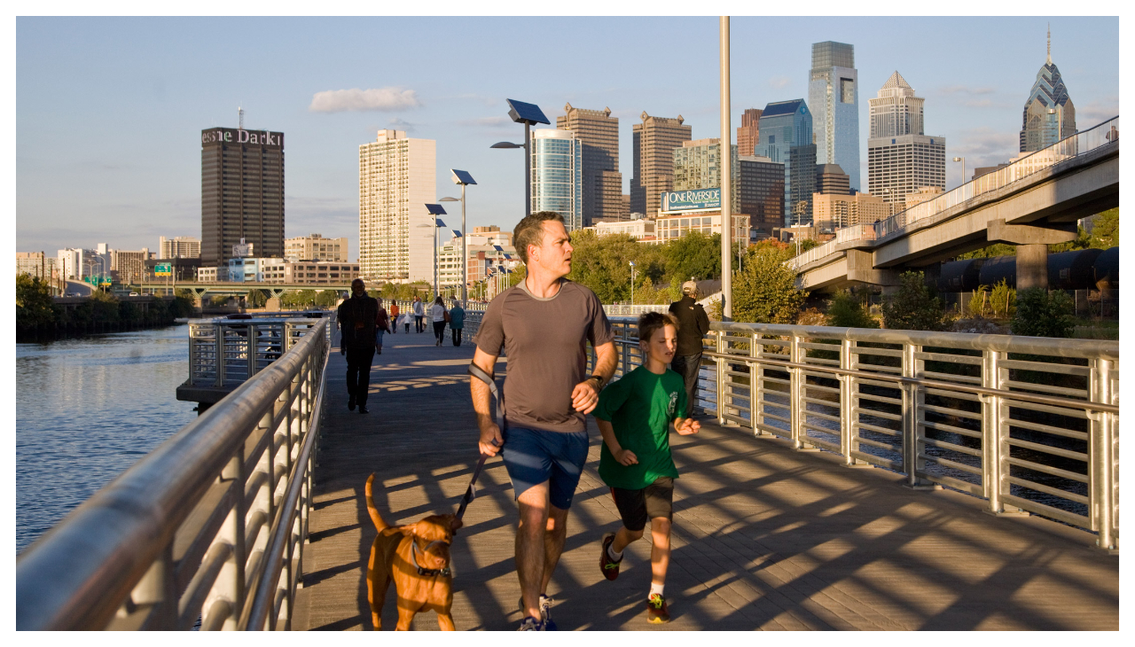 A walkable manmade path by the Schuylkill river. On it, a man walks beside his dog and a young child on the path.