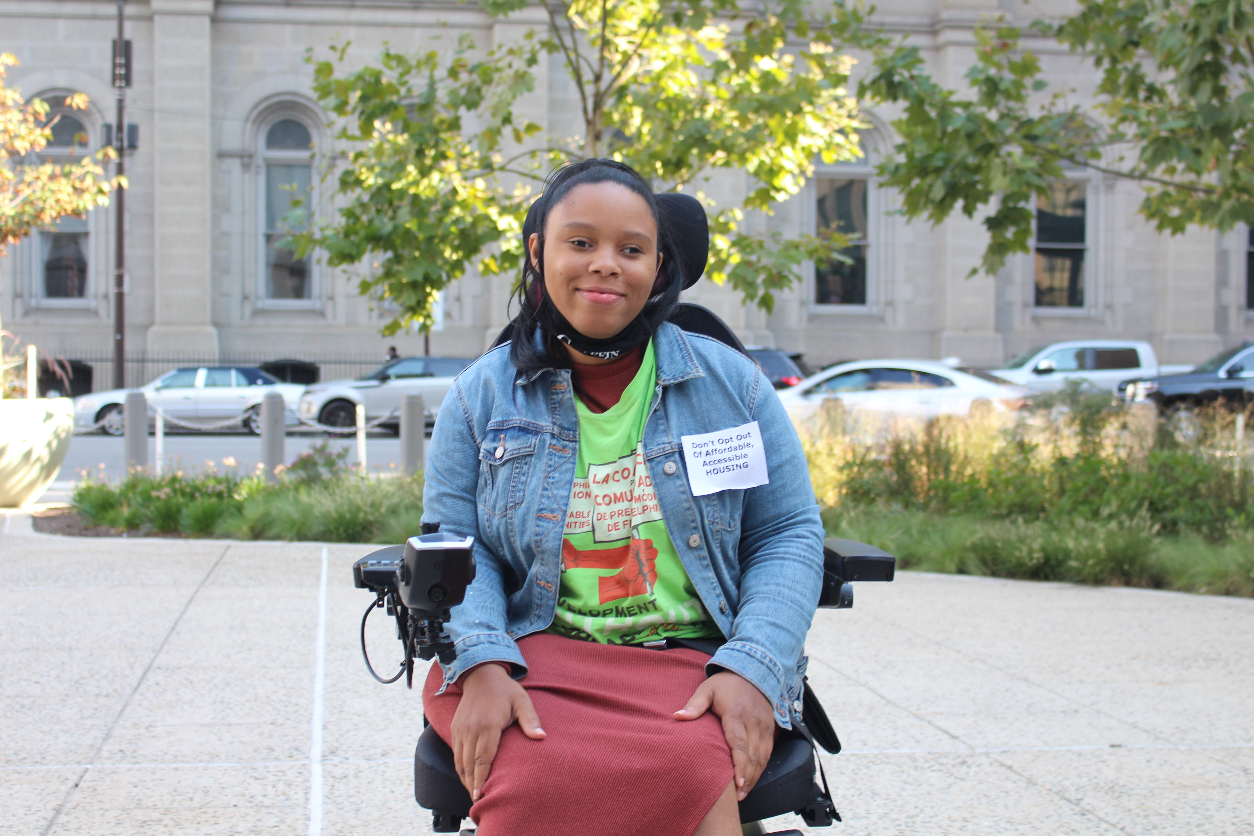 Domonique Howell, a Black woman sitting in a motorized wheelchair. She is wearing a paper badge that reads "Don't opt out of affordable, accessible housing"